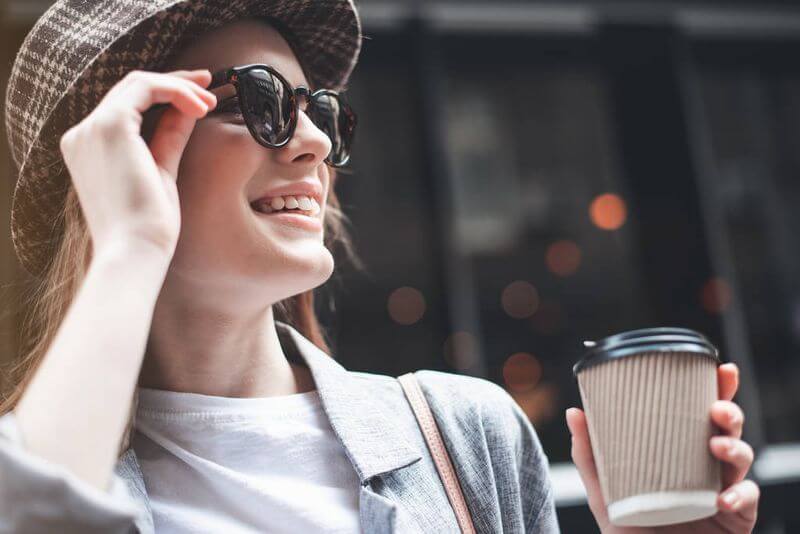 Fiatal nő napszemüvegét igazítja az utcán, kezében kávéval
